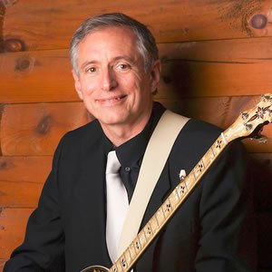 Rich Stillman in a dark shirt and white tie - you can see the neck of his banjo and the background is a wooden wall.