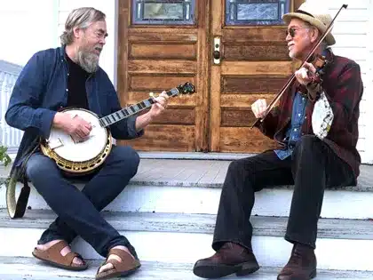 Tony Trishka sitting with a banjo next to Bruce Molsky with a fiddle.