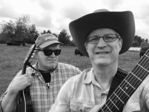 black and white photo of 2 men with a rustic scene in the background. One is holding at guitar and wearing a cowboy hat while the over is in sunglasses with a mandolin.