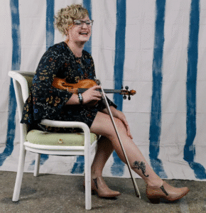 A woman sits on a chair in from of a striped backdrop, holding a fiddle and smiling.