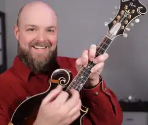 Nate, a smiling man bald with a healthy beard and mustache playing a mandolin with a strange chord fingered on the mandolin.
