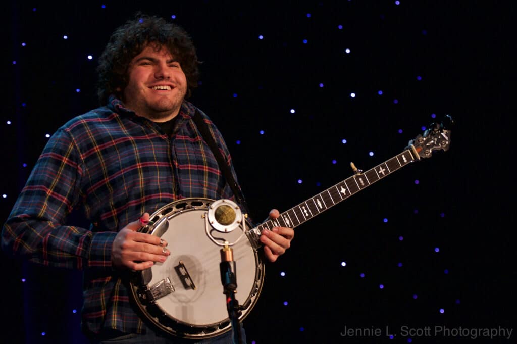 A portrait of Gabe Hirshfeld, smiling and holding his banjo with a starry backdrop at Joe Val Bluegrass Festival.