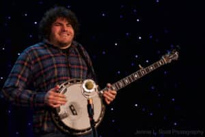 A portrait of Gabe Hirshfeld, smiling and holding his banjo with a starry backdrop at Joe Val Bluegrass Festival.