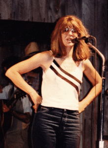 Photo of Nancy Talbot, a red-haired woman with sleeveless arms akimbo speaking into a mic.