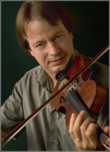 Man playing a fiddle and looking at the camera. He has brown hair and a dark khaki shirt.