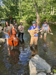 Stillwater Band members standing in a stream in the woods, holding instruments.